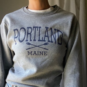 Puede incluir: Una sudadera gris con una impresión azul marino que dice "PORTLAND MAINE" con dos remos cruzados debajo del texto.