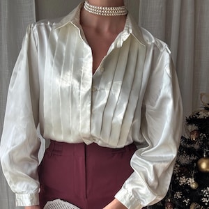 Pode incluir: Uma blusa de cetim creme com pregas verticais e mangas compridas, combinada com calças bordô. Um colar gargantilha de pérolas e uma clutch prateada com contas completam o look. Adequado para ocasiões formais.