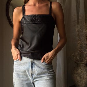 May include: Black satin camisole top with a square neckline and thin straps. The top features pleated detailing across the bust. The model is wearing light wash denim jeans. The background includes a neutral-toned curtain and a decorative vase.