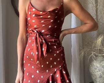 May include: A rust-coloured dress with white polka dots. The dress has a cowl neckline, spaghetti straps, and a wrap-style skirt with a ruffled hem. A matching fabric belt ties at the waist, defining the silhouette.