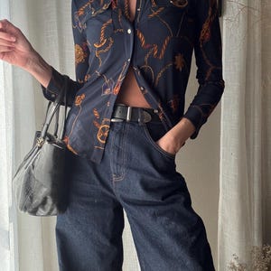 Può includere: Una camicia blu navy con un motivo a catena dorata e marrone, abbinata a jeans di denim scuro e una cintura nera. Una borsa nera è tenuta nella mano sinistra.