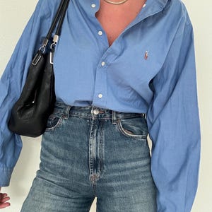 May include: A light blue button-down shirt with a small logo and high-waisted blue jeans. A black leather shoulder bag hangs on the left side. A gold-colored necklace is visible.
