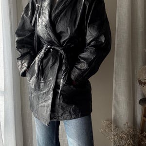 Op de afbeelding: Zwarte leren jas met een wikkelontwerp en een strikceintuur. De jas heeft een gewatteerd patroon en wordt gecombineerd met een blauwe jeans. De algehele stijl is vintage en chique.