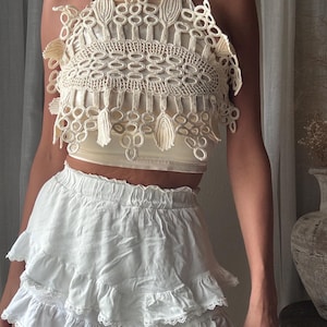 May include: A cream-colored sleeveless top featuring an intricate, raised macrame-style overlay with circular and leaf-like patterns, worn over a solid cream camisole. Paired with a white, tiered mini skirt with ruffled edges and lace trim.