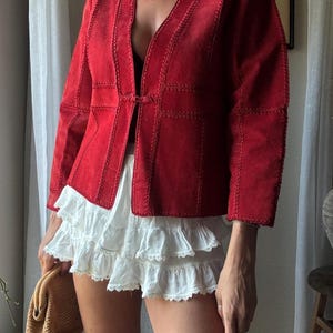 May include: A vibrant red suede jacket featuring a V-neckline, long sleeves, and decorative whipstitch detailing along the seams. The jacket has a single hook-and-eye closure at the center front. It is paired with a white tiered ruffled mini skirt.