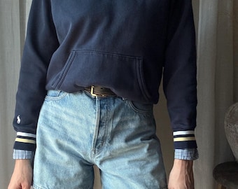 May include: Navy blue V-neck sweatshirt with a white, yellow, and navy striped collar and cuffs. The sweatshirt has a front pocket and a white embroidered logo. Worn with light wash denim shorts and a button-down shirt.