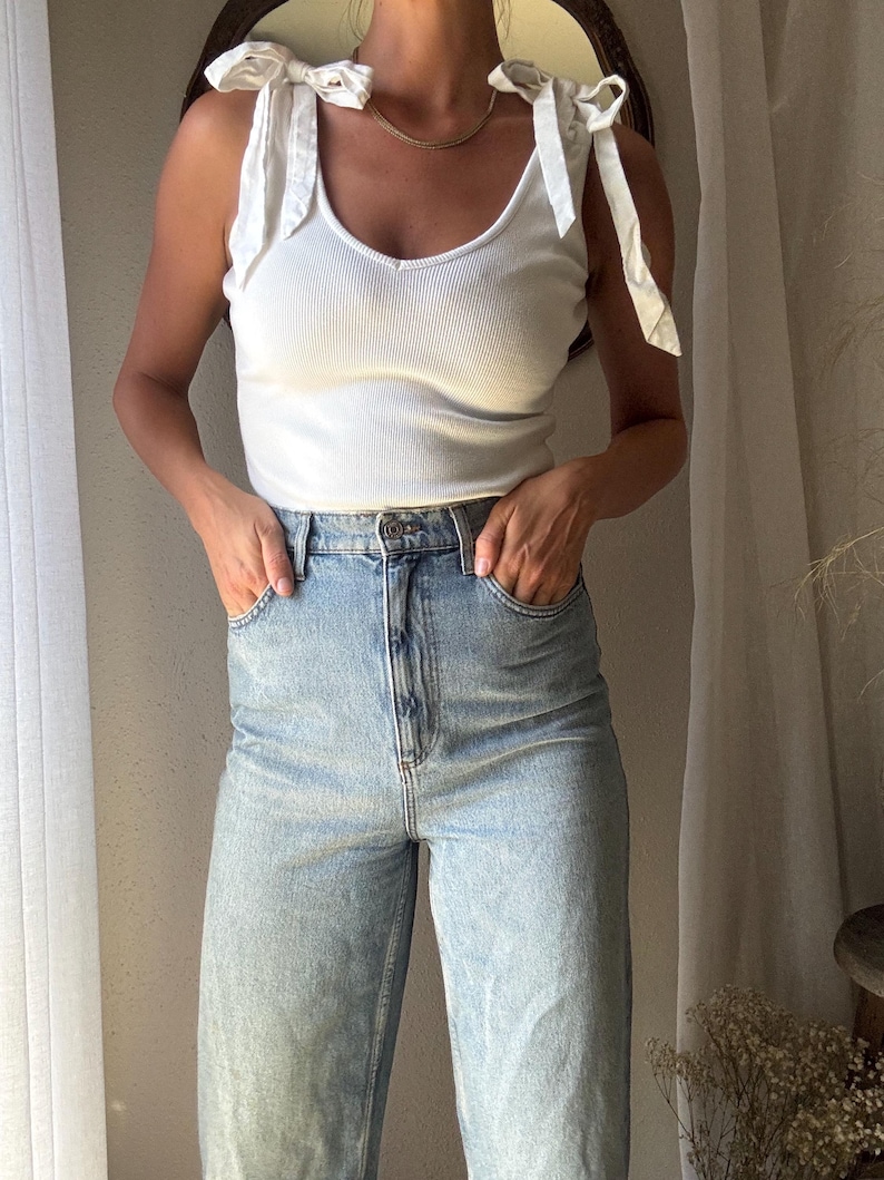 May include: A white ribbed tank top with tie straps and a V-neck is paired with light blue denim jeans. The jeans have a high waist and a relaxed fit. The outfit is simple and casual, suitable for everyday wear.