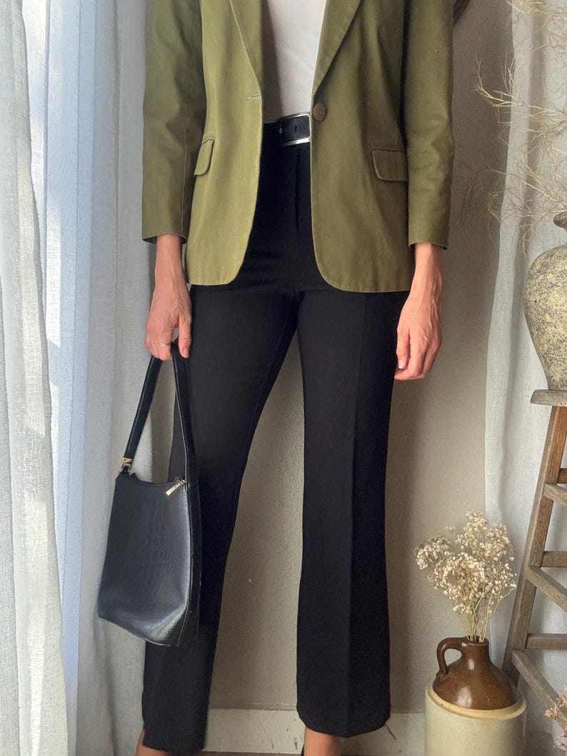 Puede incluir: Una mujer lleva una chaqueta verde sobre una camisa blanca y pantalones negros. Lleva un bolso de mano de cuero negro.