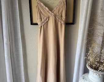 May include: A beige midi-length slip dress featuring thin spaghetti straps, a V-neckline, and vertical seam detailing. The neckline and the hem are adorned with a decorative scalloped lace trim in a matching beige tone, creating a cohesive look.