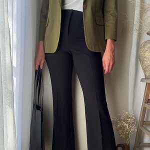 Puede incluir: Una mujer lleva una chaqueta verde sobre una camiseta blanca y pantalones negros acampanados. Lleva un bolso tote negro.