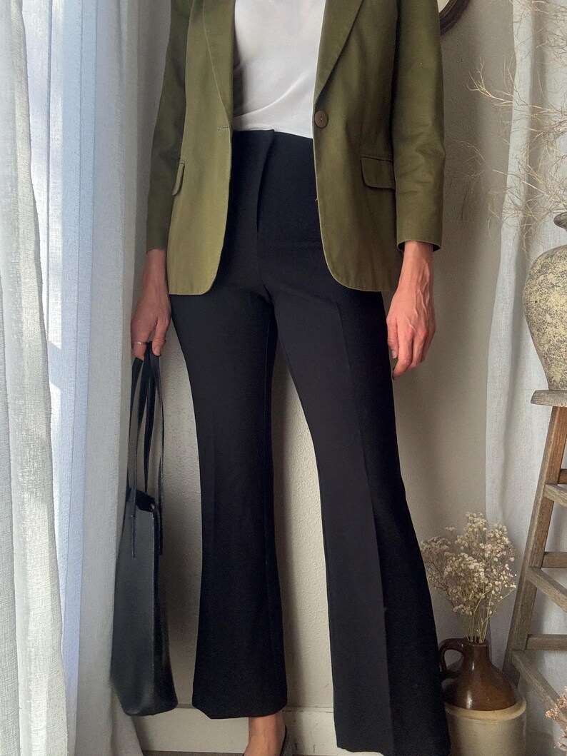 Puede incluir: Una mujer lleva una chaqueta verde sobre una camiseta blanca y pantalones negros acampanados. Lleva un bolso tote negro.