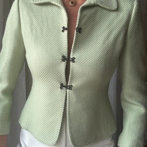 Puede incluir: Una chaqueta de color verde claro con textura acanalada diagonal, que presenta un cuello en punta, mangas tres cuartos y tres cierres de gancho negros en la parte delantera. Los bordes tienen costuras oscuras visibles a lo largo de las uniones.
