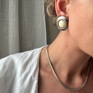May include: A silver-toned clip-on earring with a large, round, off-white center stone. The earring has a textured, hammered metal border. A gold chain necklace with a small pendant is also visible.