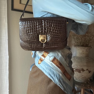 Könnte beinhalten: Eine braune, rechteckige Handtasche mit krokodilartiger Textur und einem goldfarbenen Verschluss. Die Tasche hat einen schwarzen Riemen und wird hochgehalten. Die Tasche ist im Vintage-Stil.