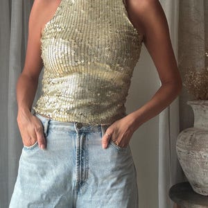 Puede incluir: Un top sin mangas de lentejuelas doradas con cuello alto, combinado con pantalones vaqueros de mezclilla azul claro. El top está cubierto de pequeñas lentejuelas metálicas doradas reflectantes que crean una superficie texturizada.