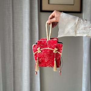 Könnte beinhalten: Eine kleine rote Stoffhandtasche mit goldenem Blumenmuster, einem einzelnen Tragegriff und zwei dekorativen goldfarbenen Quasten, die an der Vorderseite hängen. Die Tasche ist mit goldener Paspelierung und zwei kleinen runden Knoten verziert.