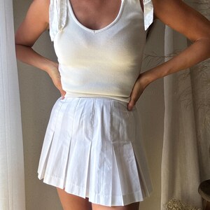 May include: A white ribbed tank top with bow tie straps and a white pleated mini skirt. The top has a V-neck and the skirt has a classic tennis skirt design. The outfit is a monochromatic ensemble.