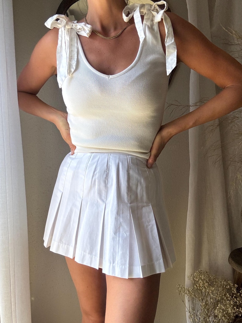 May include: A white ribbed tank top with bow tie straps and a white pleated mini skirt. The top has a V-neck and the skirt has a classic tennis skirt design. The outfit is a monochromatic ensemble.