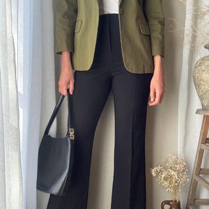 Puede incluir: Una mujer lleva una chaqueta verde sobre una camisa blanca y pantalones negros acampanados. Lleva un bolso negro.