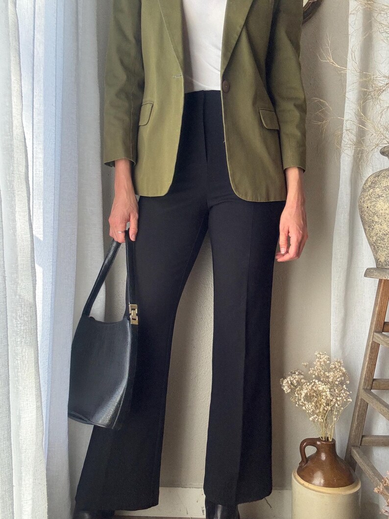 Puede incluir: Una mujer lleva una chaqueta verde sobre una camisa blanca y pantalones negros acampanados. Lleva un bolso negro.