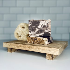 May include: Two bars of handmade soap on a wooden tray. The soap is brown and white with a marbled pattern. The tray is made of light brown wood and has a simple design.