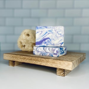 May include: Two bars of handmade soap on a wooden tray. The soap is white with purple swirls. The tray is made of light brown wood and has a simple design.