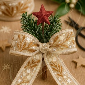 May include: A festive Christmas bow with a red glitter star, pine sprigs, and a tan ribbon featuring a white floral pattern. The bow is tied and mounted on a cinnamon stick, perfect for holiday decor.
