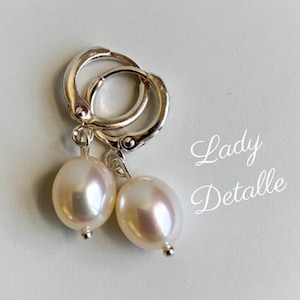 Pode incluir: Brincos de argola de prata com uma pérola branca pendurada em cada argola. Os brincos estão sobre um fundo branco com o texto "Lady Detalle" em preto.