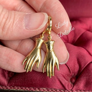 K&ouml;nnte beinhalten: Goldfarbene Ohrringe mit einem Paar H&auml;nden mit einem detaillierten Design. Die Ohrringe h&auml;ngen an einem Ring.