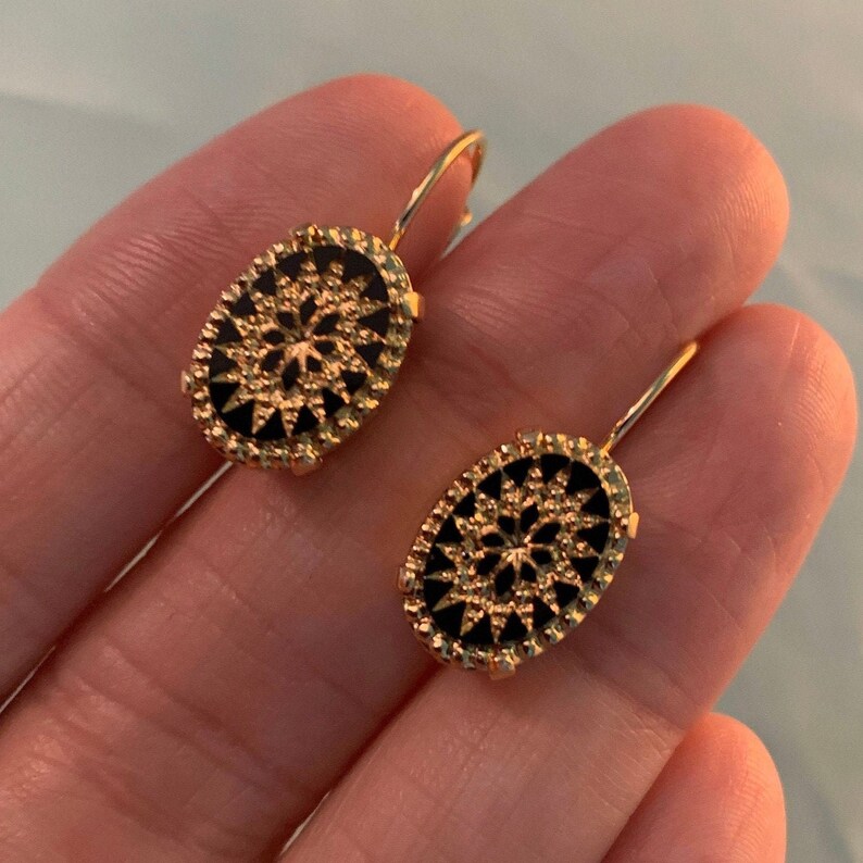 May include: A pair of gold-tone earrings with black enamel oval-shaped drop pendants. Each pendant features a detailed starburst design.