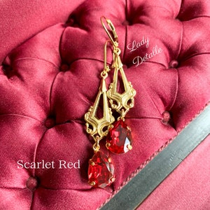 May include: Gold dangle earrings with a red teardrop-shaped crystal. The earrings feature a delicate, ornate design. The text "Lady Detalle" is visible in the background.