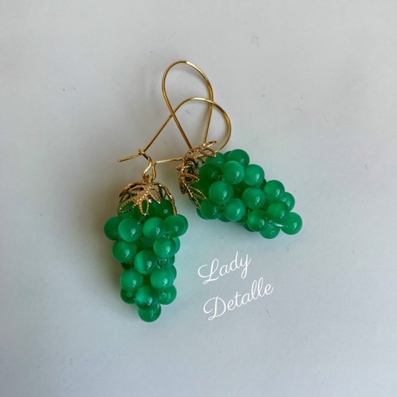 May include: A pair of green grape earrings with gold hooks. The grapes are made of glass beads and are clustered together to form a bunch.
