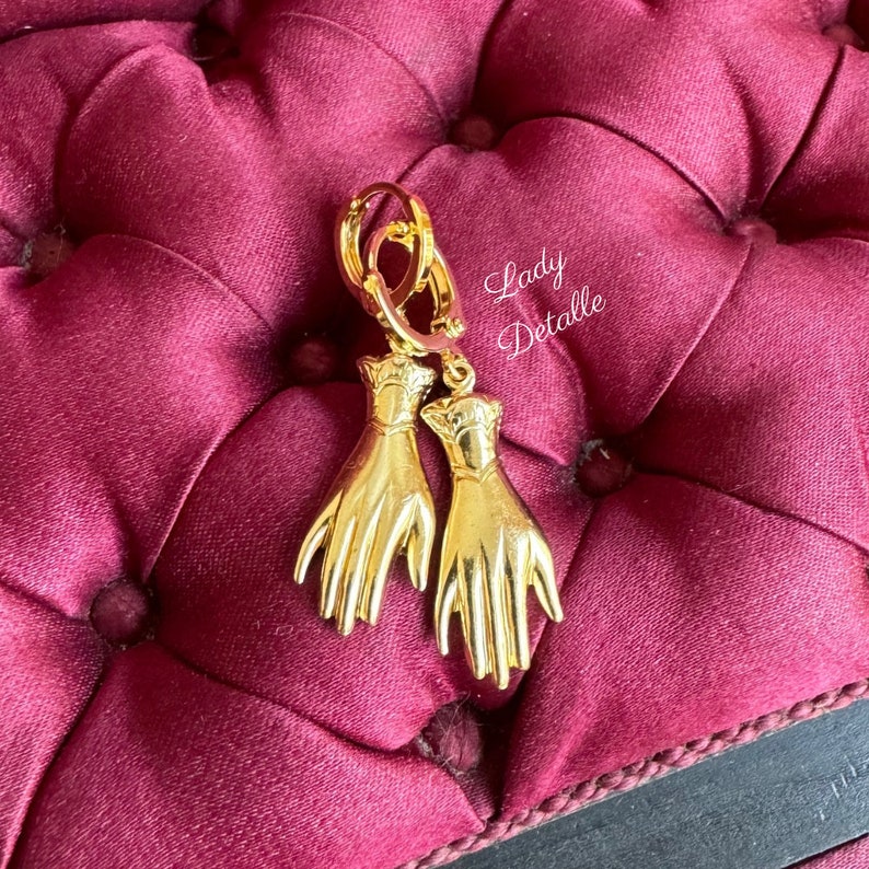 K&ouml;nnte beinhalten: Goldene Creolen mit einem baumelnden Handdesign. Die H&auml;nde sind detailliert und haben einen Vintage-Look.