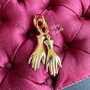 K&ouml;nnte beinhalten: Goldfarbene Ohrringe mit einem Handdesign. Die Ohrringe h&auml;ngen an einem Kreis.