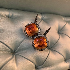May include: A pair of orange-colored, round earrings with a crystal border and dark metal hooks. The earrings are set against a light blue, tufted background, creating a contrast of colors and textures. The earrings have a warm, radiant glow.