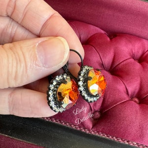 May include: A pair of leverback earrings featuring large, faceted orange and yellow crystals. Each crystal is surrounded by a halo of small, clear crystals set in a black metal frame. The earrings are held against a burgundy velvet surface.