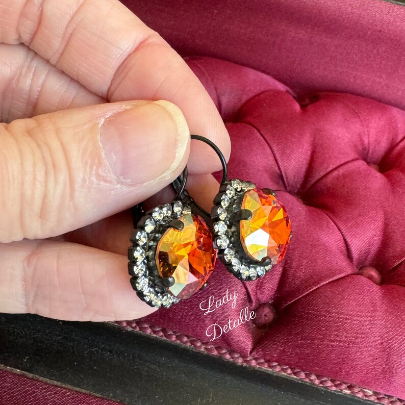 May include: A pair of leverback earrings featuring large, faceted orange and yellow crystals. Each crystal is surrounded by a halo of small, clear crystals set in a black metal frame. The earrings are held against a burgundy velvet surface.