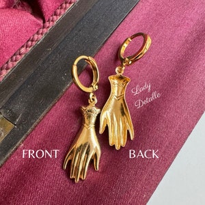 K&ouml;nnte beinhalten: Goldene Creolen mit einem baumelnden Hand-Anh&auml;nger. Der Anh&auml;nger ist mit einer strukturierten Oberfl&auml;che und einem erhabenen Design auf der R&uuml;ckseite verziert. Die Ohrringe sind auf der R&uuml;ckseite mit "Lady Detalle" beschriftet.