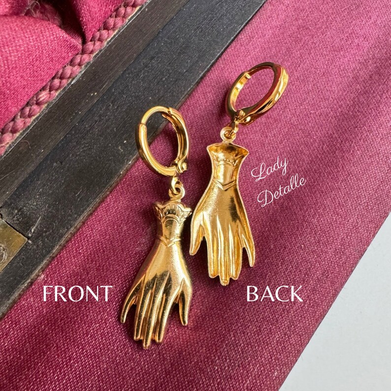 K&ouml;nnte beinhalten: Goldene Creolen mit einem baumelnden Hand-Anh&auml;nger. Der Anh&auml;nger ist mit einer strukturierten Oberfl&auml;che und einem erhabenen Design auf der R&uuml;ckseite verziert. Die Ohrringe sind auf der R&uuml;ckseite mit "Lady Detalle" beschriftet.