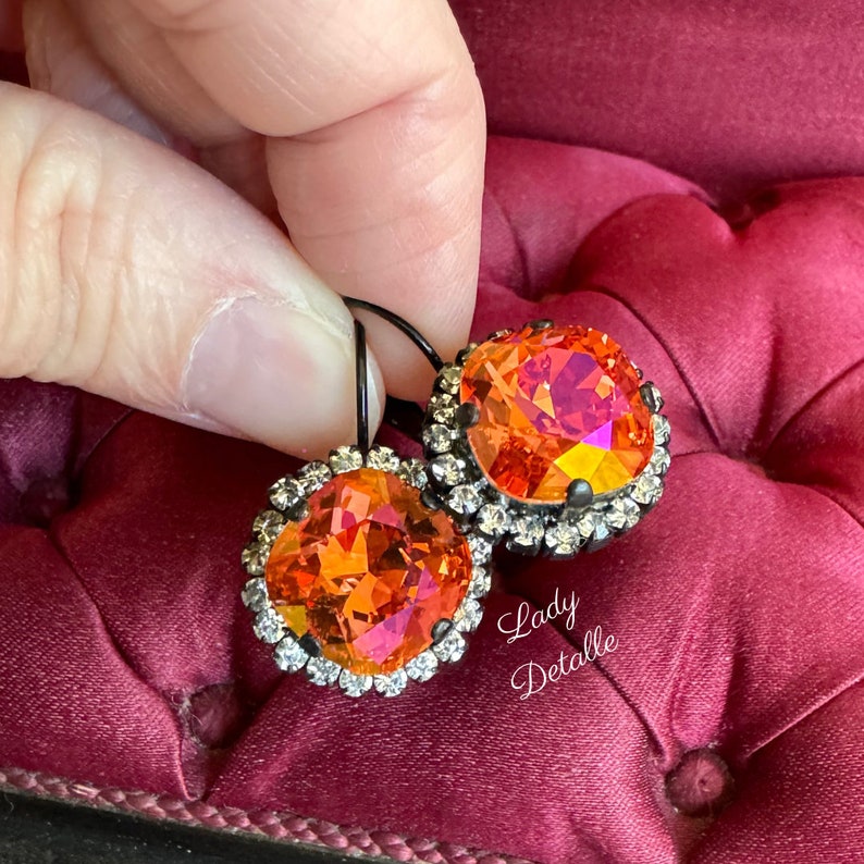 May include: A pair of orange and pink crystal earrings with a halo of small clear crystals. The earrings have black metal hooks and are set against a deep red background. The text "Lady Detalle" is in the lower right corner.