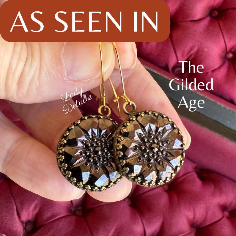 K&ouml;nnte beinhalten: Ein Paar goldfarbene Ohrringe mit einem dunkelbraunen, facettierten, knopfartigen Design. Die Ohrringe h&auml;ngen an einem Ohr einer Person und der Text "The Gilded Age" ist im Hintergrund sichtbar.