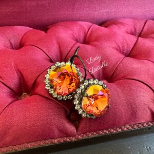 May include: A pair of orange crystal earrings with a halo of small clear crystals. The earrings have a dark metal setting and are displayed on a pink, tufted surface. The text "Lady Detalle" is visible.