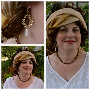 May include: A woman wearing a white blouse with a gold and brown patterned head scarf, a gold necklace with purple stones, and gold earrings with pearl drops.