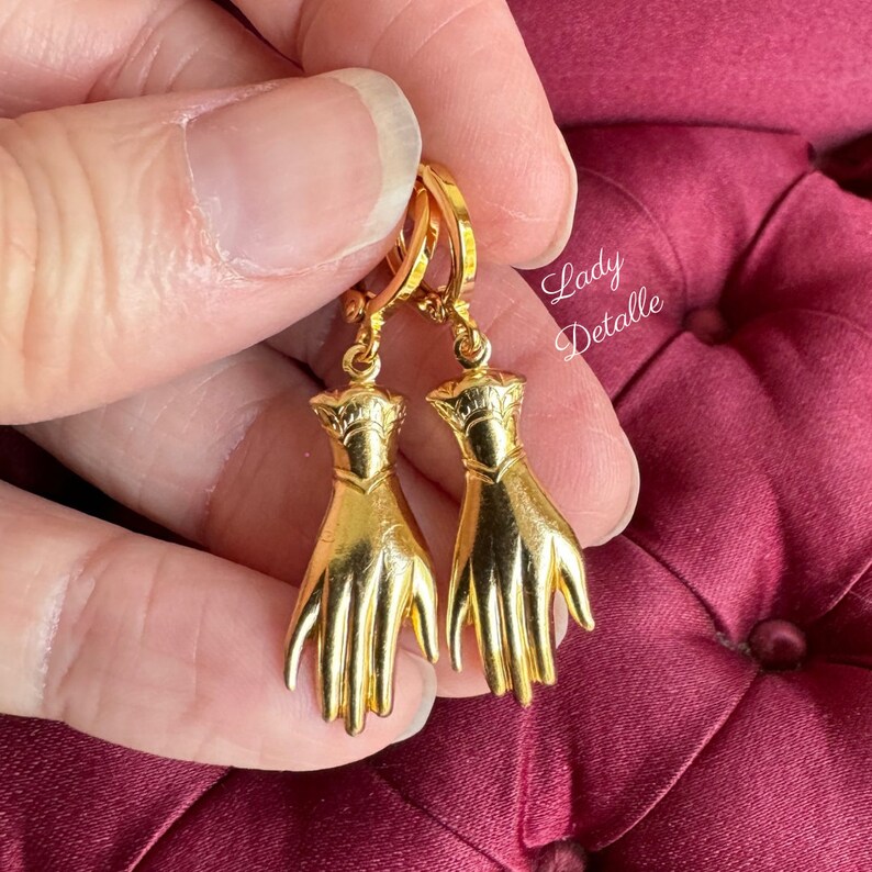 K&ouml;nnte beinhalten: Goldene Creolen mit einem baumelnden Hand-Anh&auml;nger. Die H&auml;nde sind detailliert und haben eine strukturierte Oberfl&auml;che.