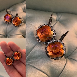 May include: A pair of square-cut, orange crystal earrings with a dark metal setting and a halo of small, clear crystals. The earrings are displayed on a light blue surface.