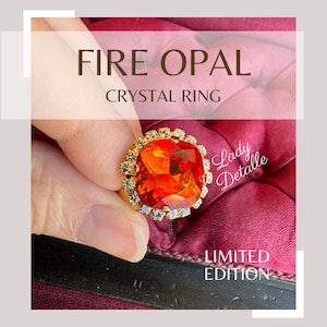 May include: A gold ring featuring a large, bright orange fire opal gemstone surrounded by smaller clear crystals. The ring is labelled "Fire Opal Crystal Ring" and "Limited Edition".