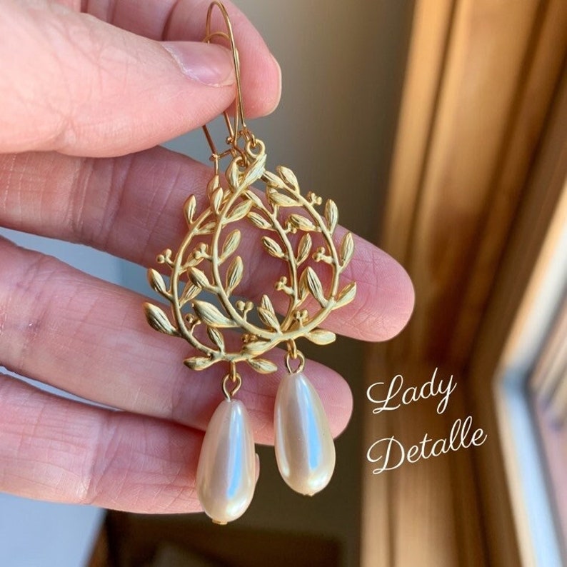 May include: Gold earrings with a delicate leaf design and two pearlescent white teardrop-shaped beads hanging from the bottom.
