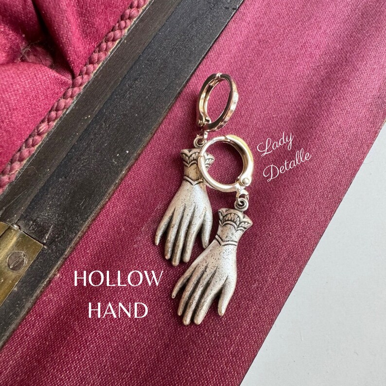 K&ouml;nnte beinhalten: Silberne Ohrringe mit einem hohlen Handdesign. Die Ohrringe h&auml;ngen an einem silbernen Kreis und haben ein zartes, detailliertes Design. Die Ohrringe sind mit "Hollow Hand" und "Lady Detalle" beschriftet.