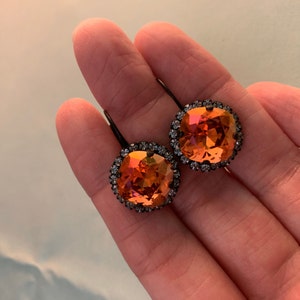May include: A pair of orange and yellow crystal earrings with a dark metal setting. Each earring features a large, square-cut crystal surrounded by smaller, clear crystals. The earrings are held by a hand.