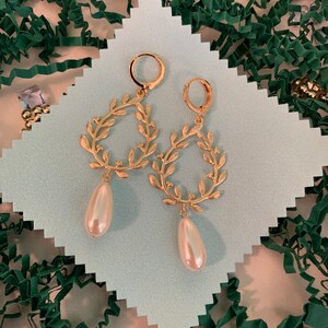 May include: Gold hoop earrings with a delicate leaf design and a dangling pearl drop. The earrings are displayed on a white background.